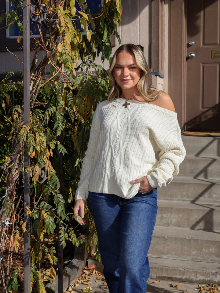 Woman wearing a white off-shoulder sweater and blue jeans standing outdoors.