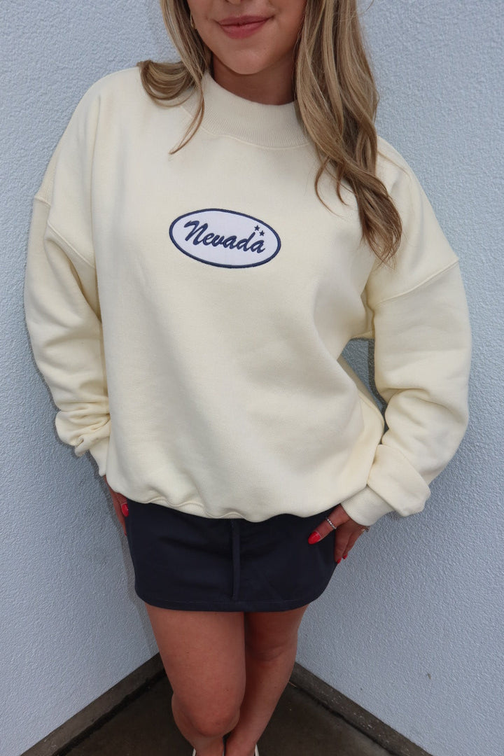 A woman wearing a cream-colored crewneck sweatshirt with an oval 'Nevada' logo on the chest, paired with a dark navy mini skirt and white Mary Jane shoes, standing against a light blue textured wall.