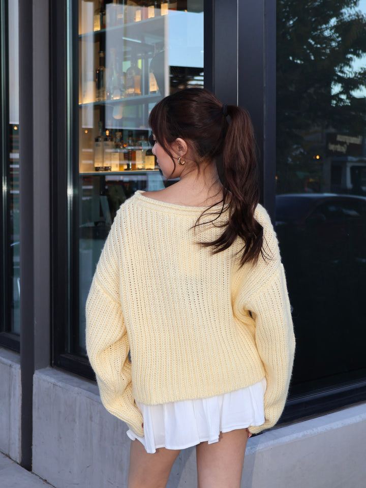 Person wearing a light yellow sweater and white shorts standing in front of a store window.