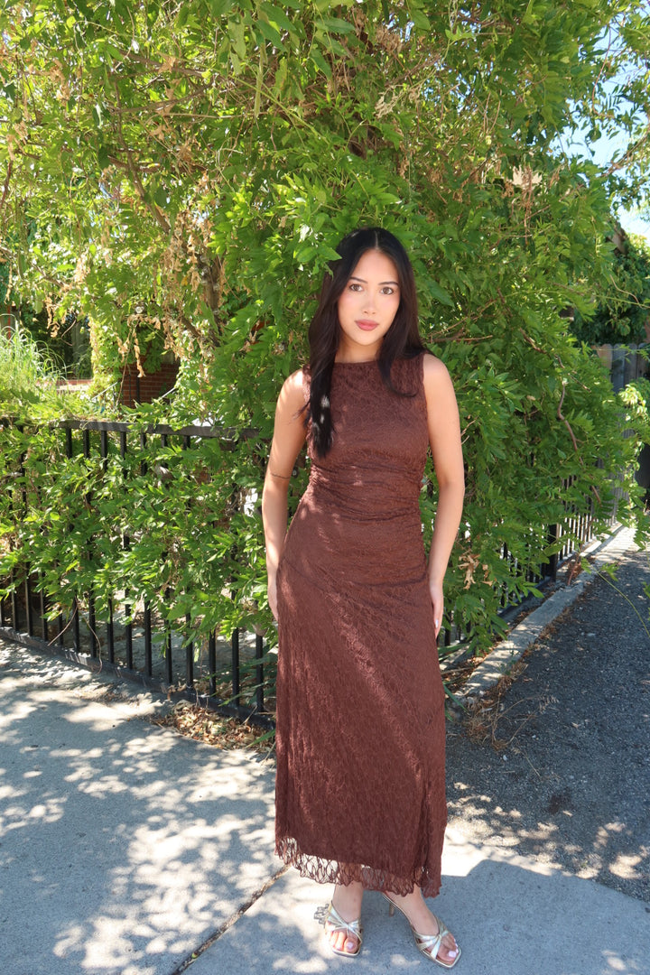 A woman wearing a sleeveless, knee-length brown lace dress stands outdoors on a sidewalk next to a black metal fence and a leafy green tree.