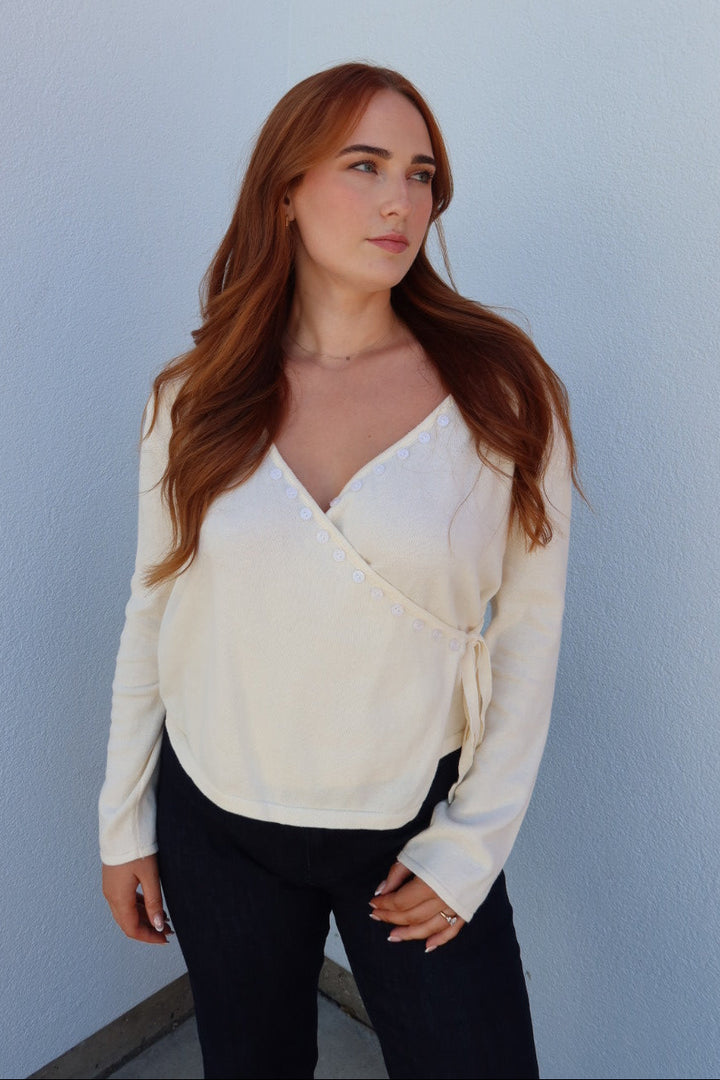 A woman with long red hair wearing a cream-colored long-sleeve wrap-style blouse and black pants, standing against a light blue-gray wall.
