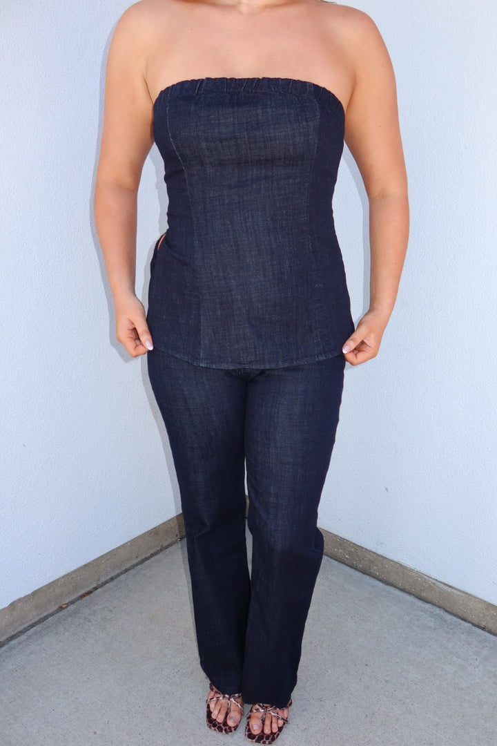 A woman modeling a dark indigo blue strapless tube top and matching dark blue jeans, standing in a corner against a light blue wall.