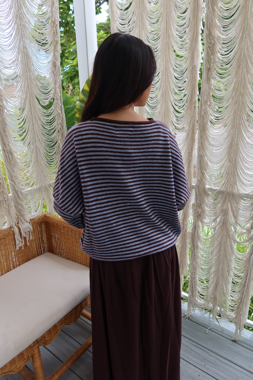 A woman viewed from behind wearing a navy and gray striped long-sleeve top and a dark brown skirt, standing on a porch in front of white macrame curtains.