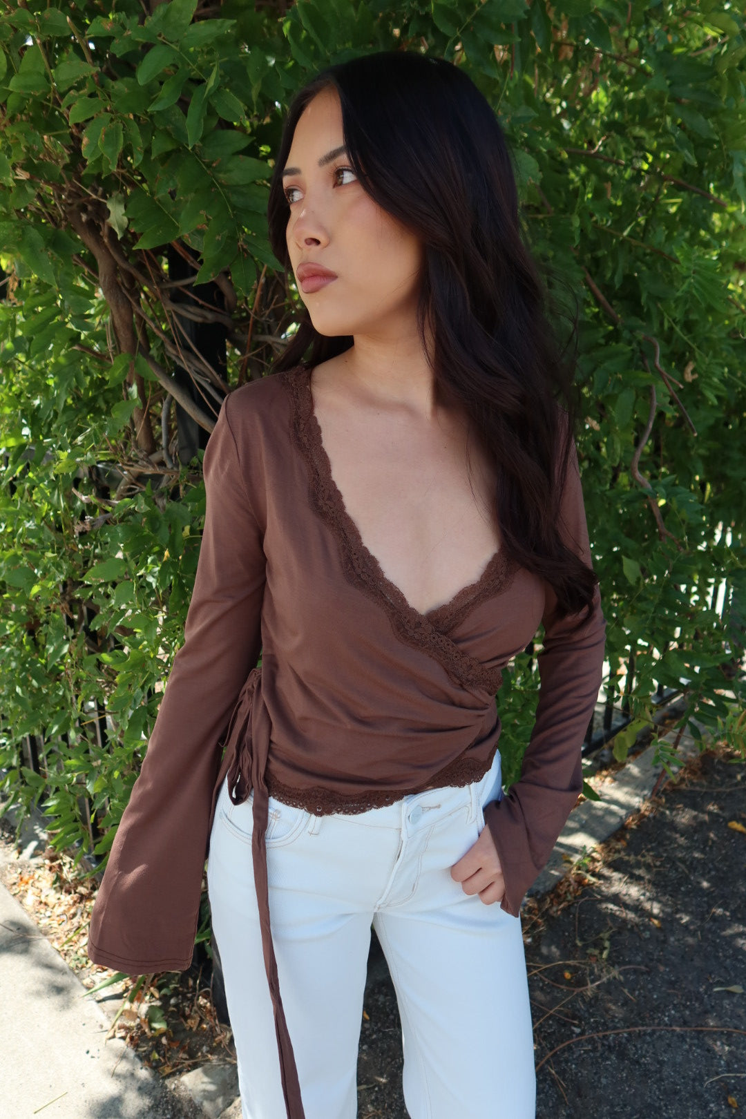 A woman wearing a brown long-sleeve wrap-style top with lace trim and white pants, standing outdoors in front of green foliage.
