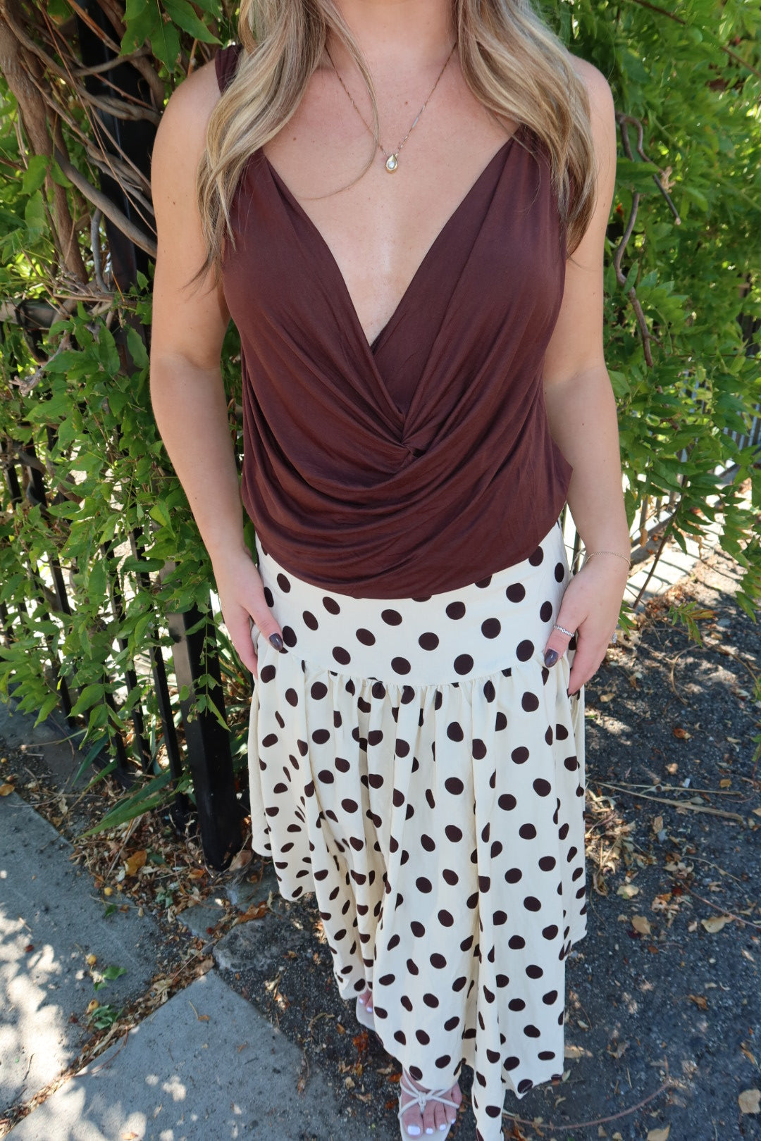A woman wearing a brown draped wrap-style tank top and a white skirt with dark brown polka dots, standing outdoors in front of green foliage.