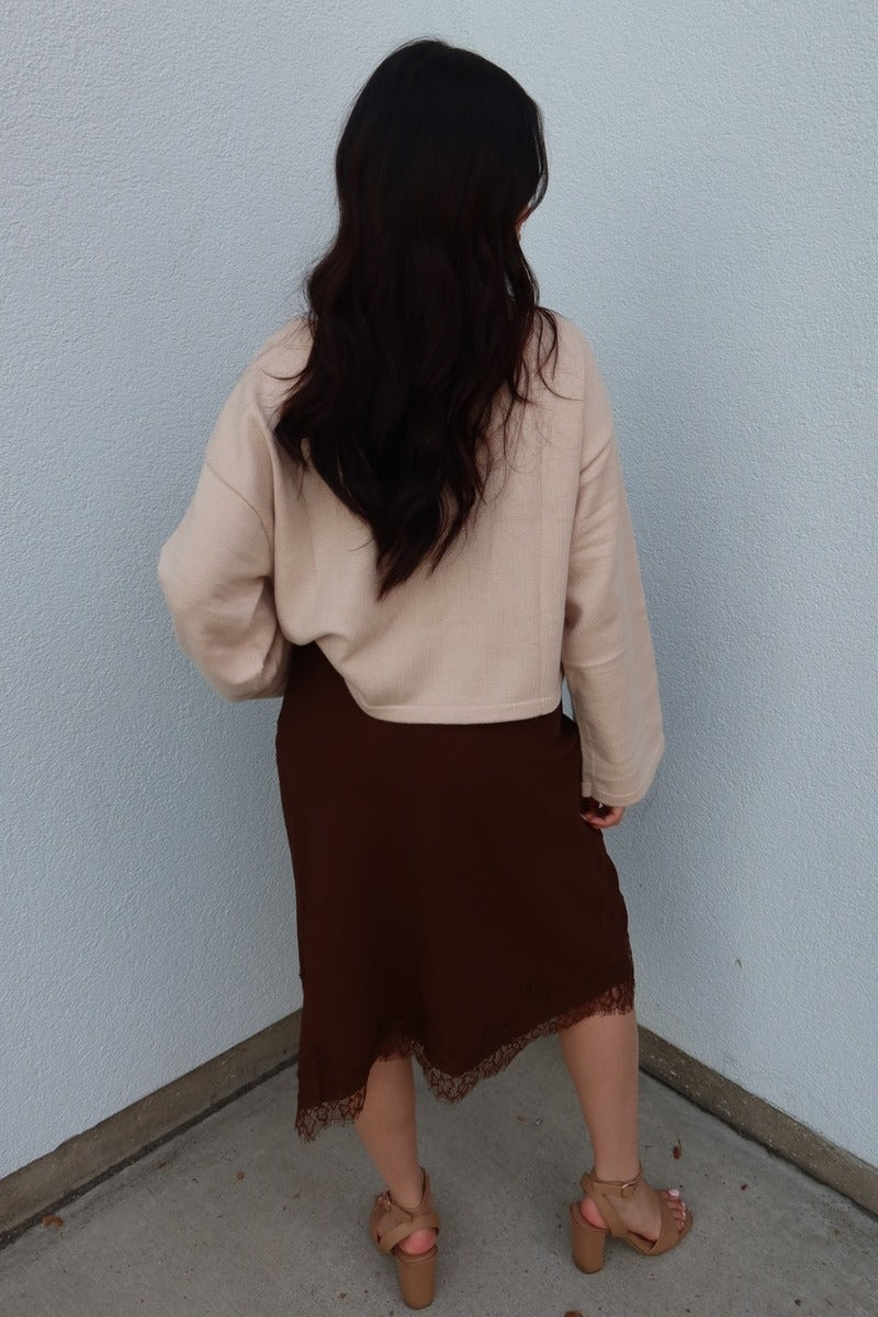 Angled back view of the brown Lucy asymmetric lace trim satin midi skirt with a cream cardigan and tan heels