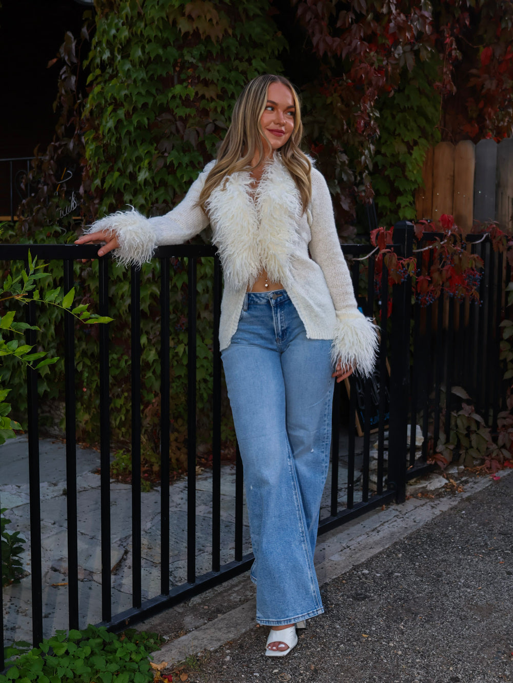 Woman wearing a white fur-trimmed jacket and blue jeans standing outdoors.