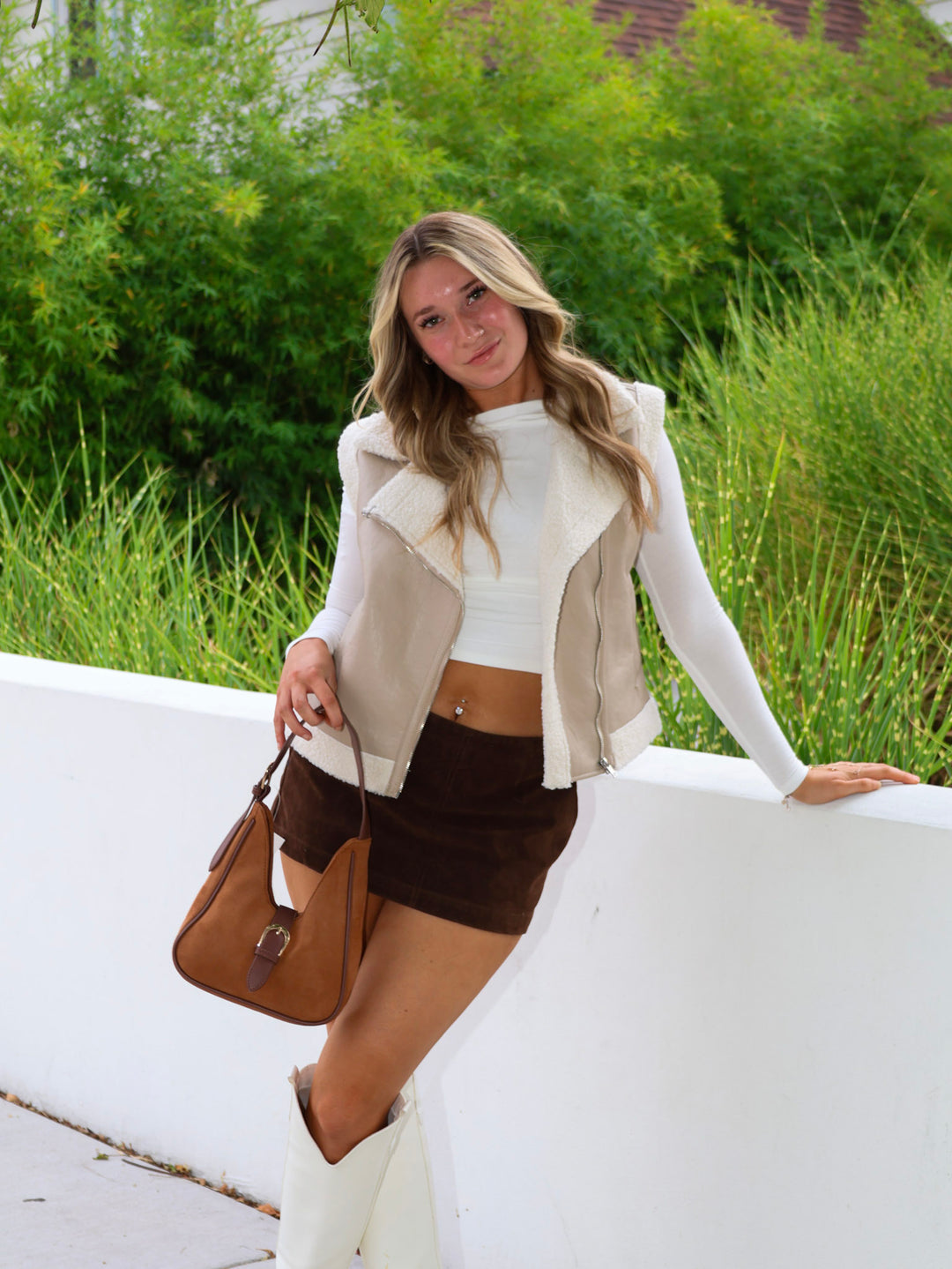 Woman in a white jacket and brown shorts holding a brown handbag outdoors.