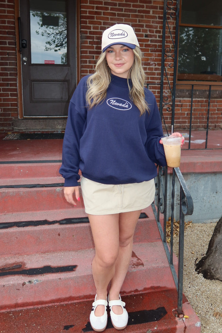 A woman stands on the red brick steps of a building wearing a navy blue sweatshirt with a white oval logo, a beige mini skirt, white shoes, and a white cap. She is holding a drink.