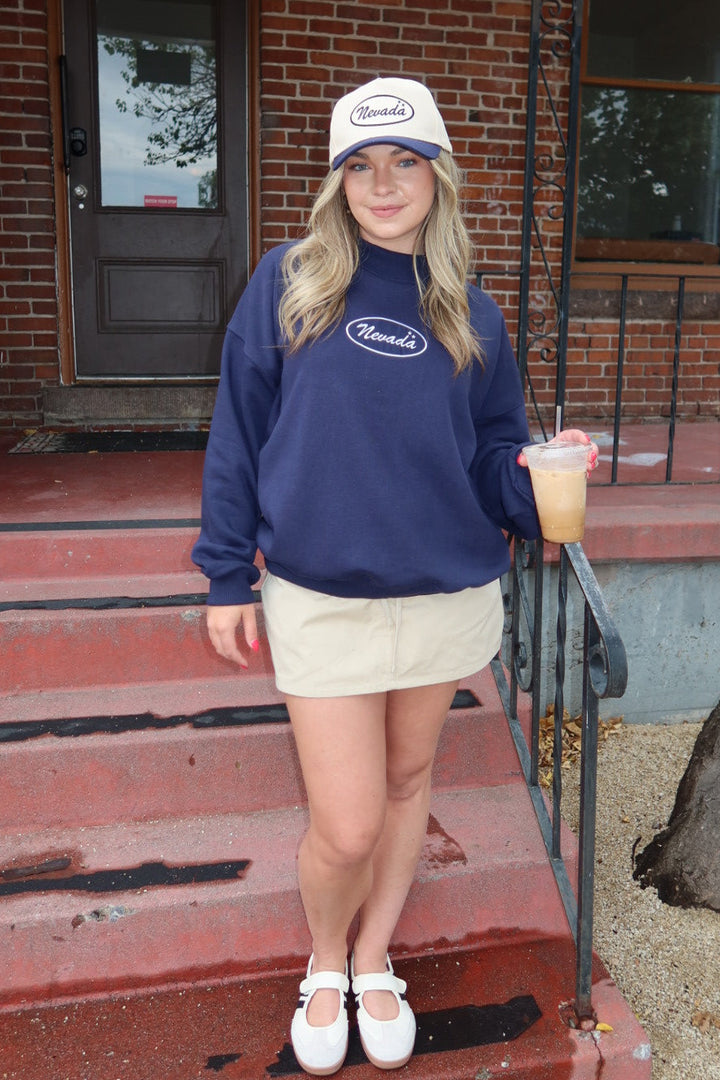 A woman stands on the red brick steps of a building wearing a navy blue sweatshirt with a white oval logo, a beige mini skirt, white shoes, and a white cap. She is holding a drink.