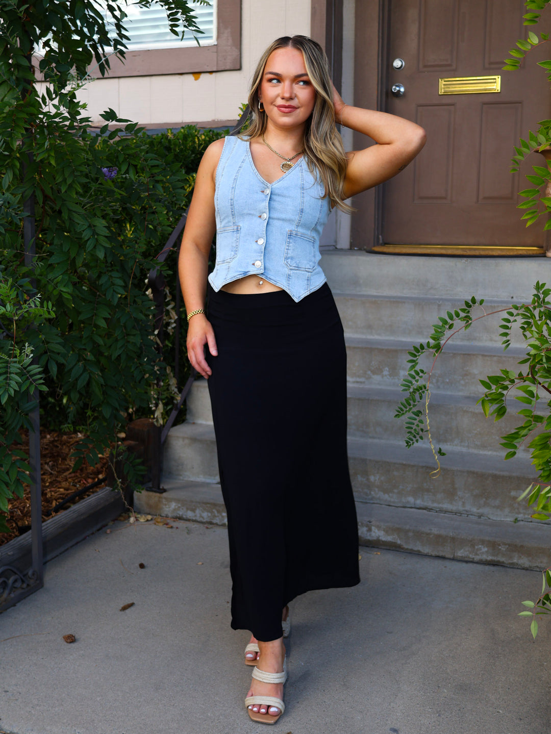 Woman wearing a denim vest and black skirt standing on steps outside a house.