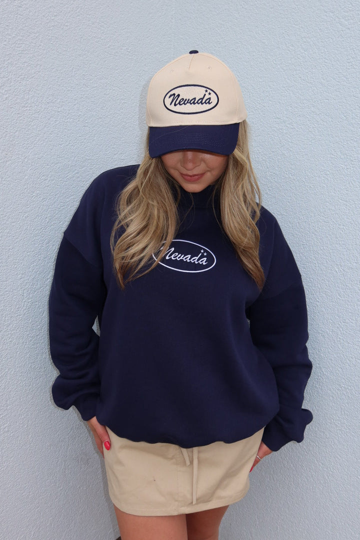 A woman wearing a navy blue sweatshirt and matching baseball cap with 'Nevada' embroidered in white, paired with a beige mini skirt, standing against a light blue textured wall.