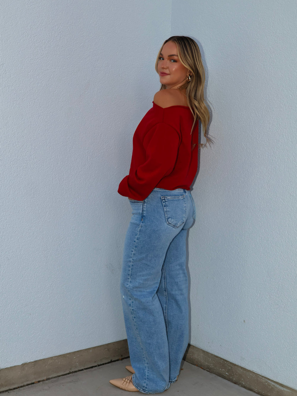 Person wearing a red top and blue jeans standing against a white wall.