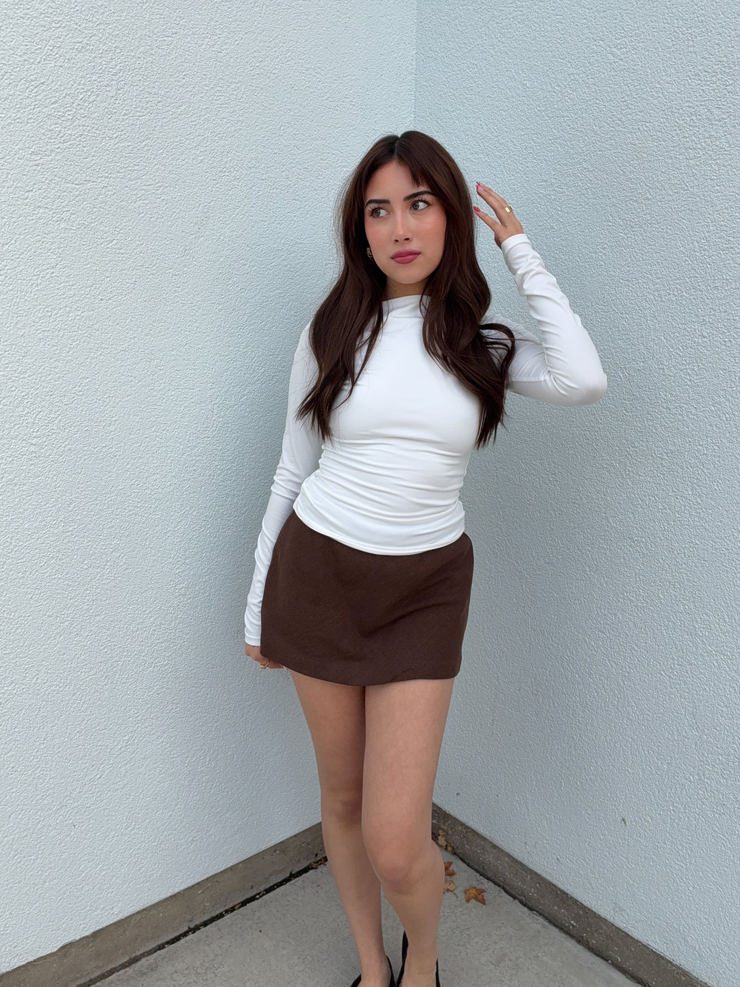 Woman wearing a white long-sleeve top and brown skirt against a light blue wall.