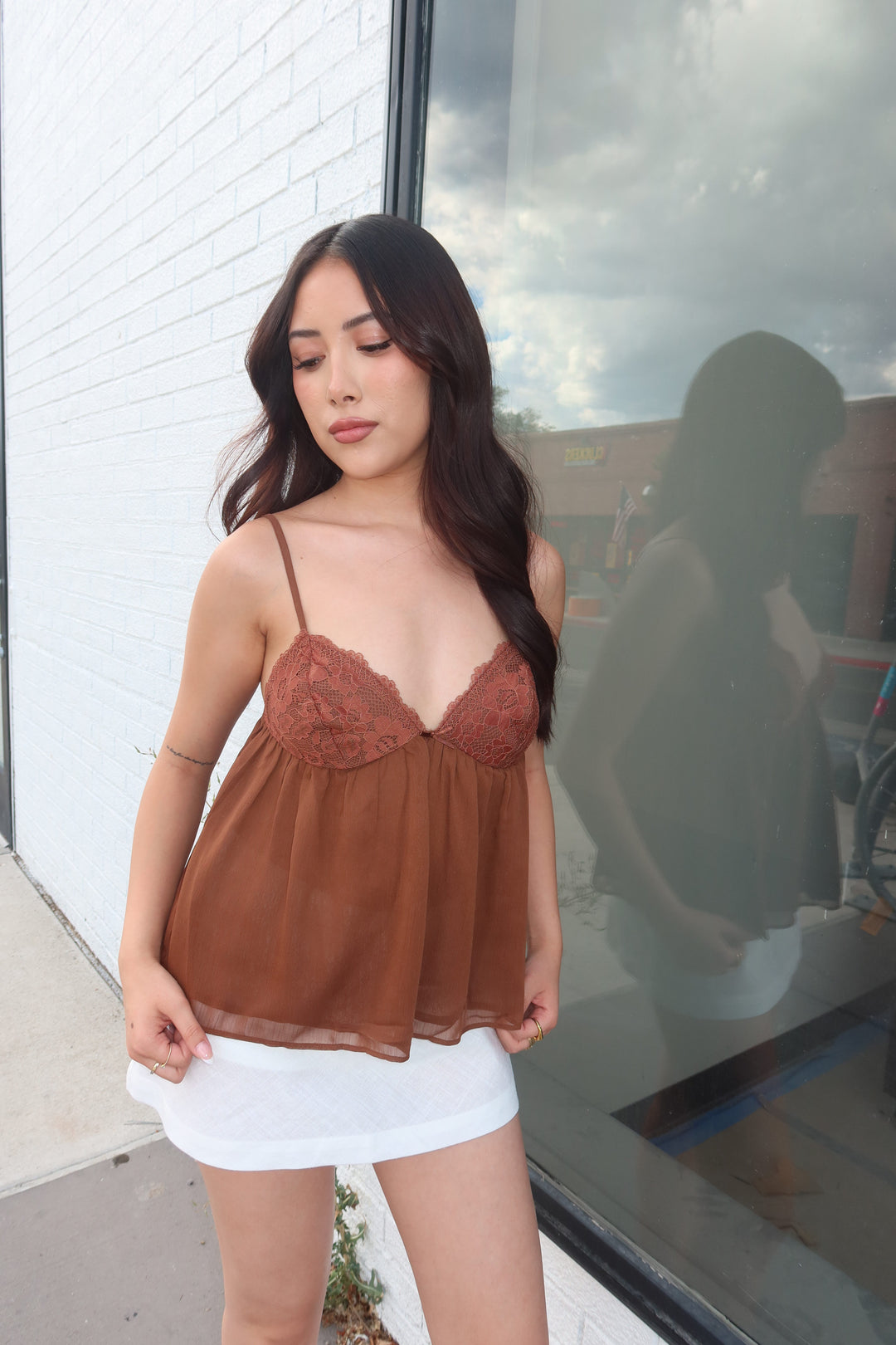 A woman wearing a brown lace-trim camisole top and a white skirt, photographed outdoors near a glass window with reflections.