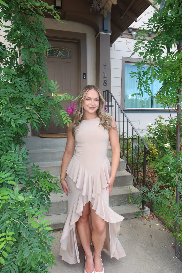 A woman wearing a sleeveless high-low dress in a light blush pink or nude beige color, standing on the concrete steps of a house with green foliage on both sides.