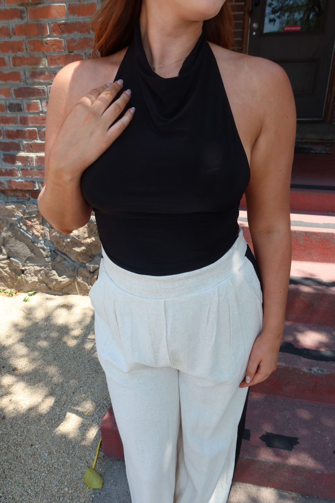 A woman wearing a black halter-neck top and white high-waisted pleated trousers, standing outdoors in front of a brick wall and stone steps.