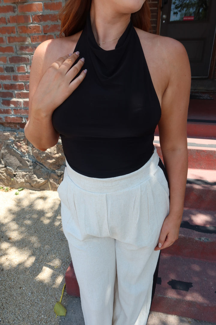 A woman wearing a black halter-neck top and white high-waisted pleated trousers, standing outdoors in front of a brick wall and stone steps.