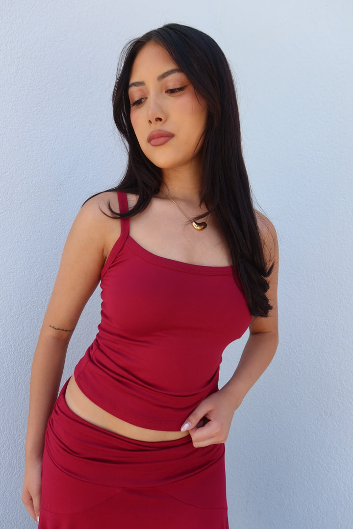 A woman with long dark hair models a matching red ribbed knit two-piece set consisting of a spaghetti-strap tank top and a ruched high-waisted skirt, set against a light blue textured wall.