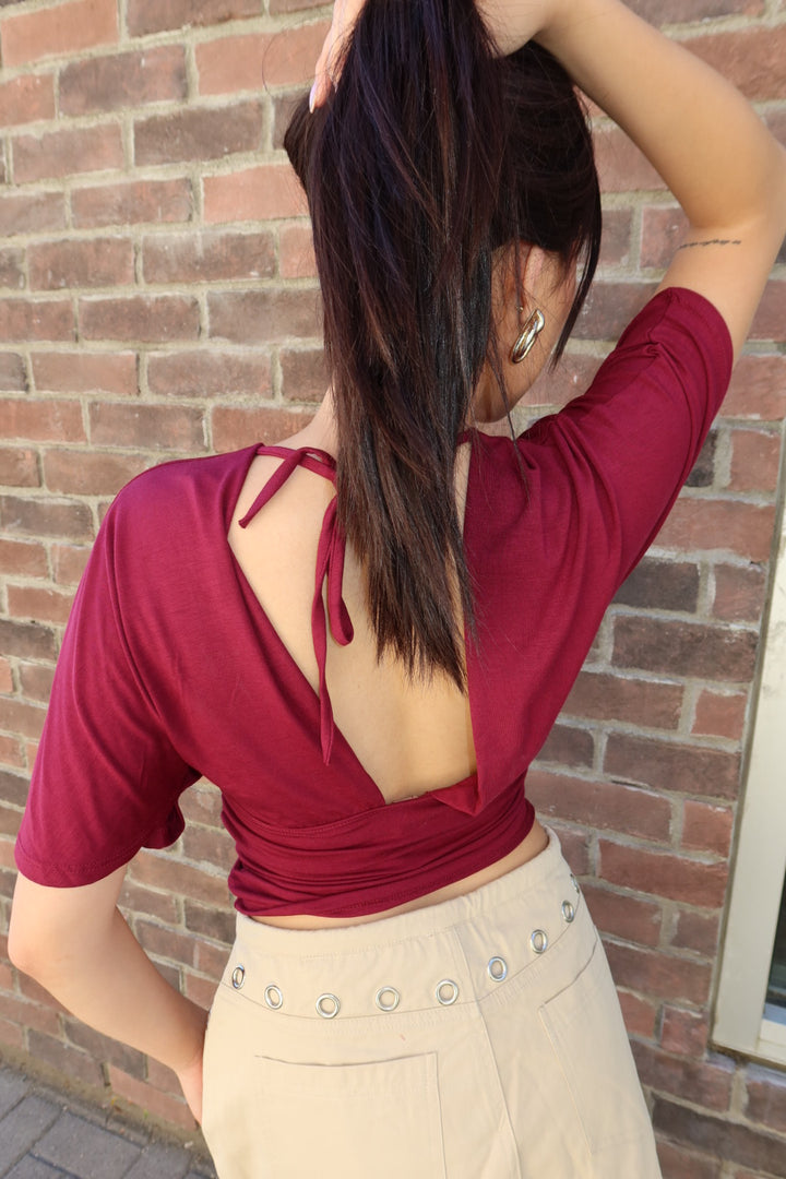 A woman shown from the back wearing a short-sleeve burgundy top with a deep open back and tie detail, paired with beige high-waisted shorts, standing against a brick wall.