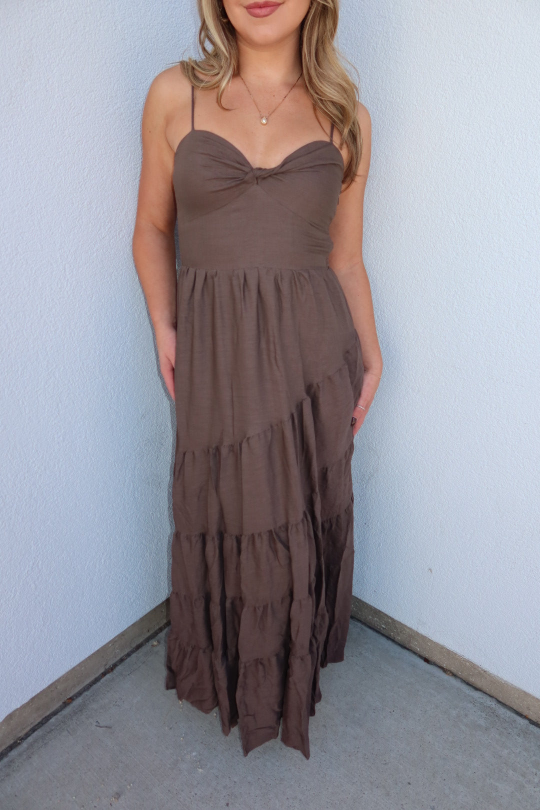 A woman wearing a brown tiered maxi dress with spaghetti straps and a sweetheart neckline, standing against a light blue textured wall.
