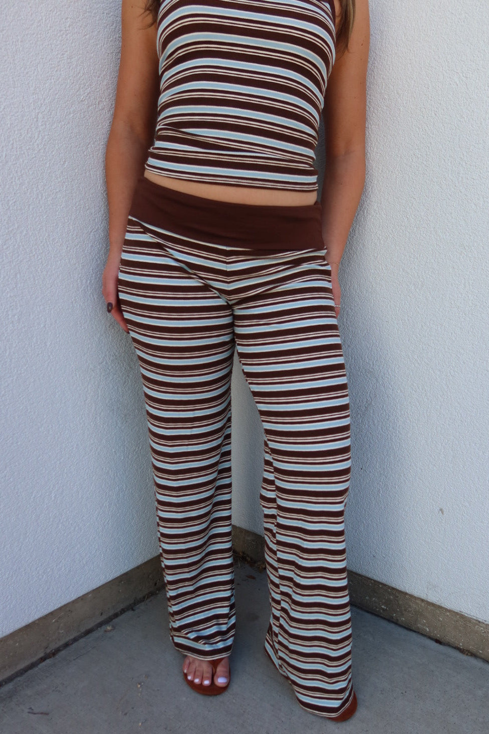A person wearing a brown, beige, and white striped matching set consisting of a sleeveless crop top and wide-leg pants, standing against a white textured wall.