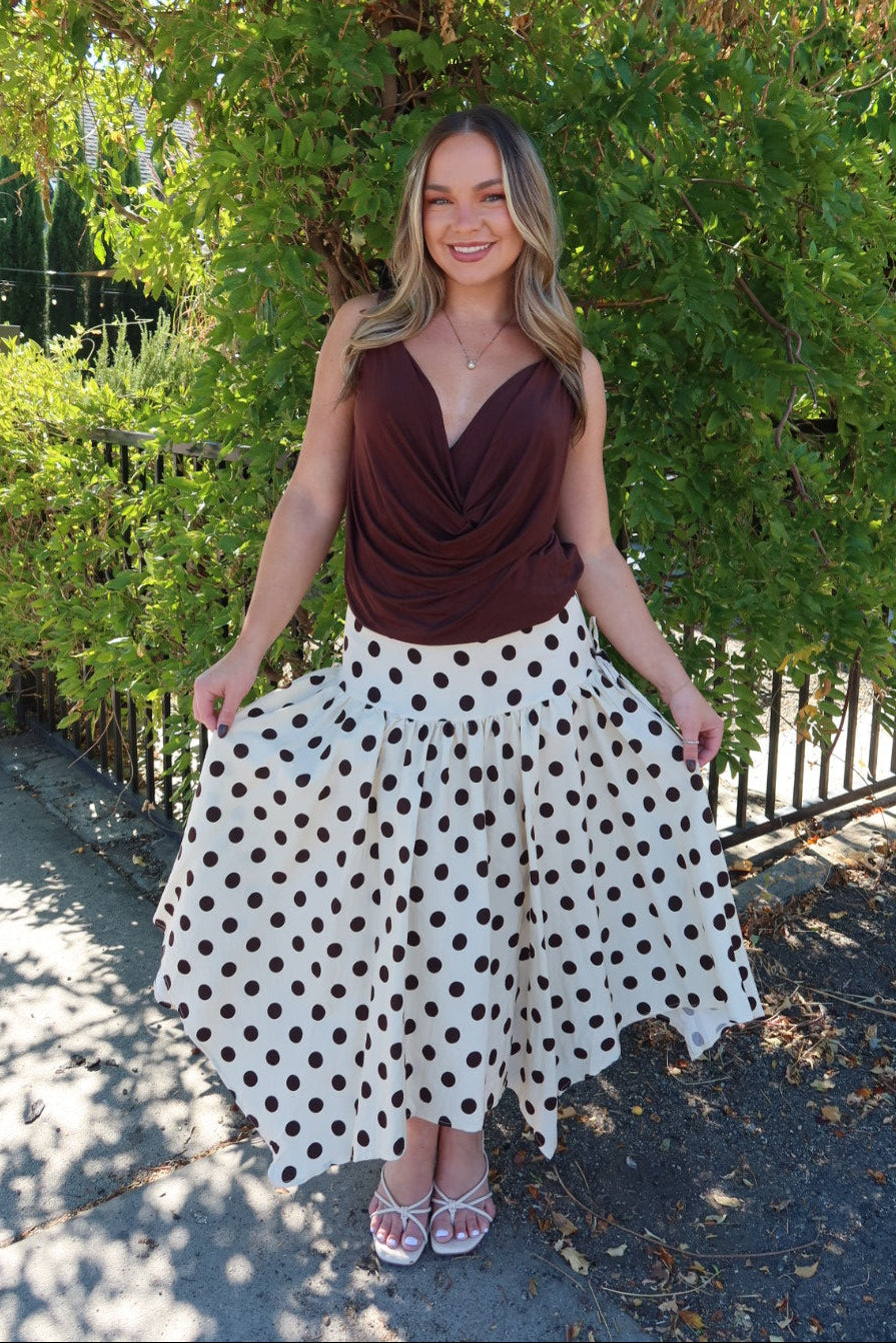 A woman stands outdoors in front of a leafy green tree, wearing a sleeveless dark brown draped top and a white midi skirt with brown polka dots. She is holding out the sides of the skirt to show its full volume.