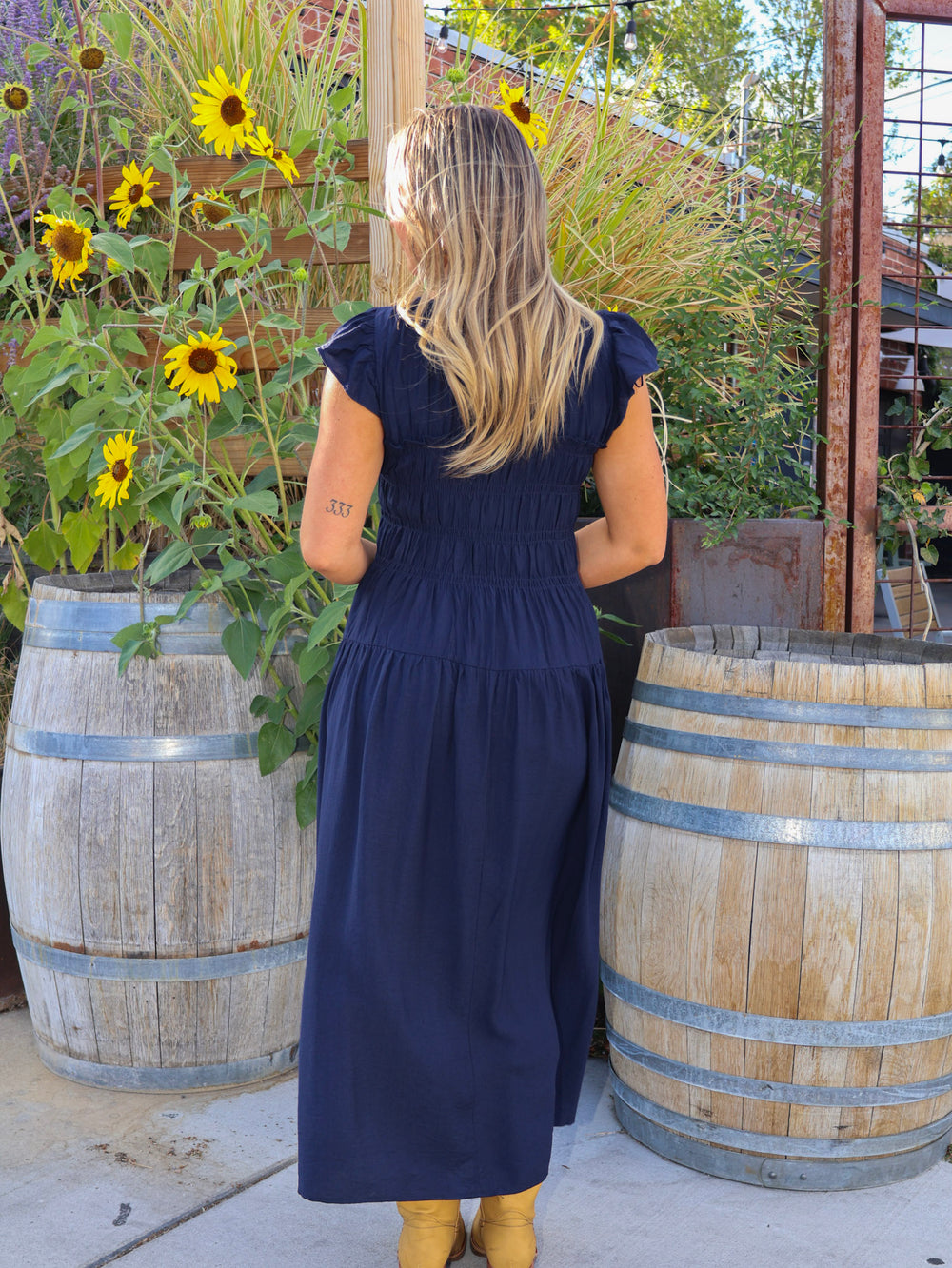 Woman in a navy dress standing in a garden with sunflowers and wooden barrels.
