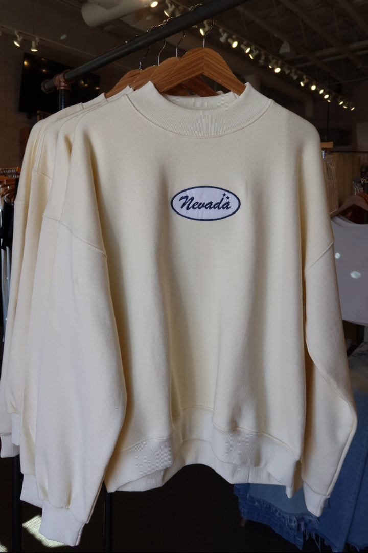 An off-white crewneck sweatshirt hanging on a wooden hanger in a retail store. The sweatshirt features an oval chest logo with the word 'Nevada' in blue script inside a white oval with a blue border.