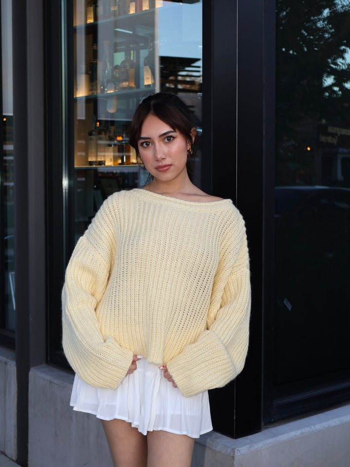 Woman wearing a light yellow sweater and white skirt standing outside a building.