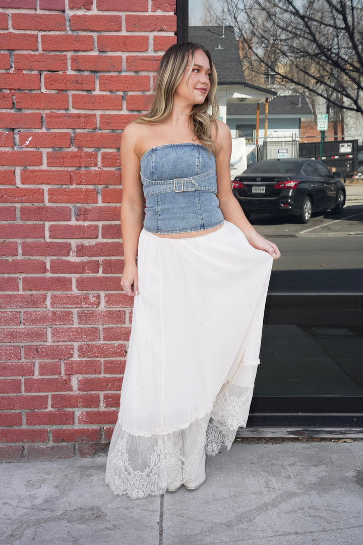 Woman in a denim top and white skirt standing against a brick wall.