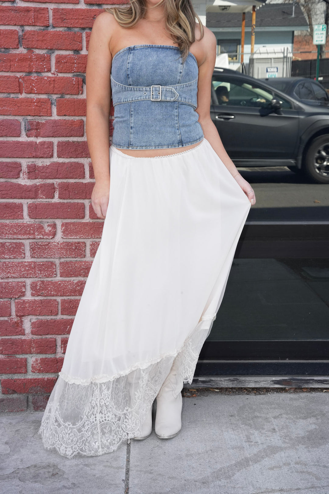 Person wearing a denim crop top and white skirt with lace hem against a brick wall.