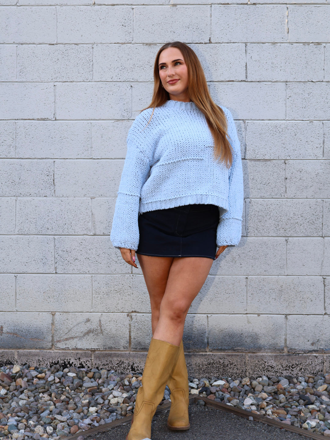 Woman wearing a light blue sweater, black skirt, and tan boots standing against a gray brick wall.