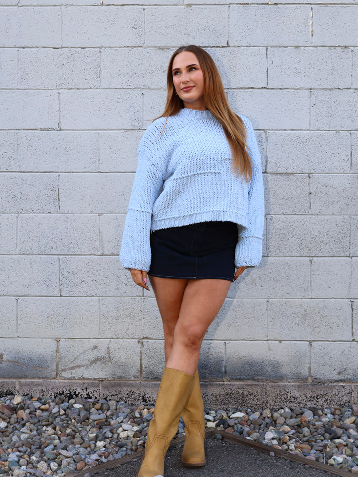Woman wearing a light blue sweater, black skirt, and tan boots standing against a gray brick wall.