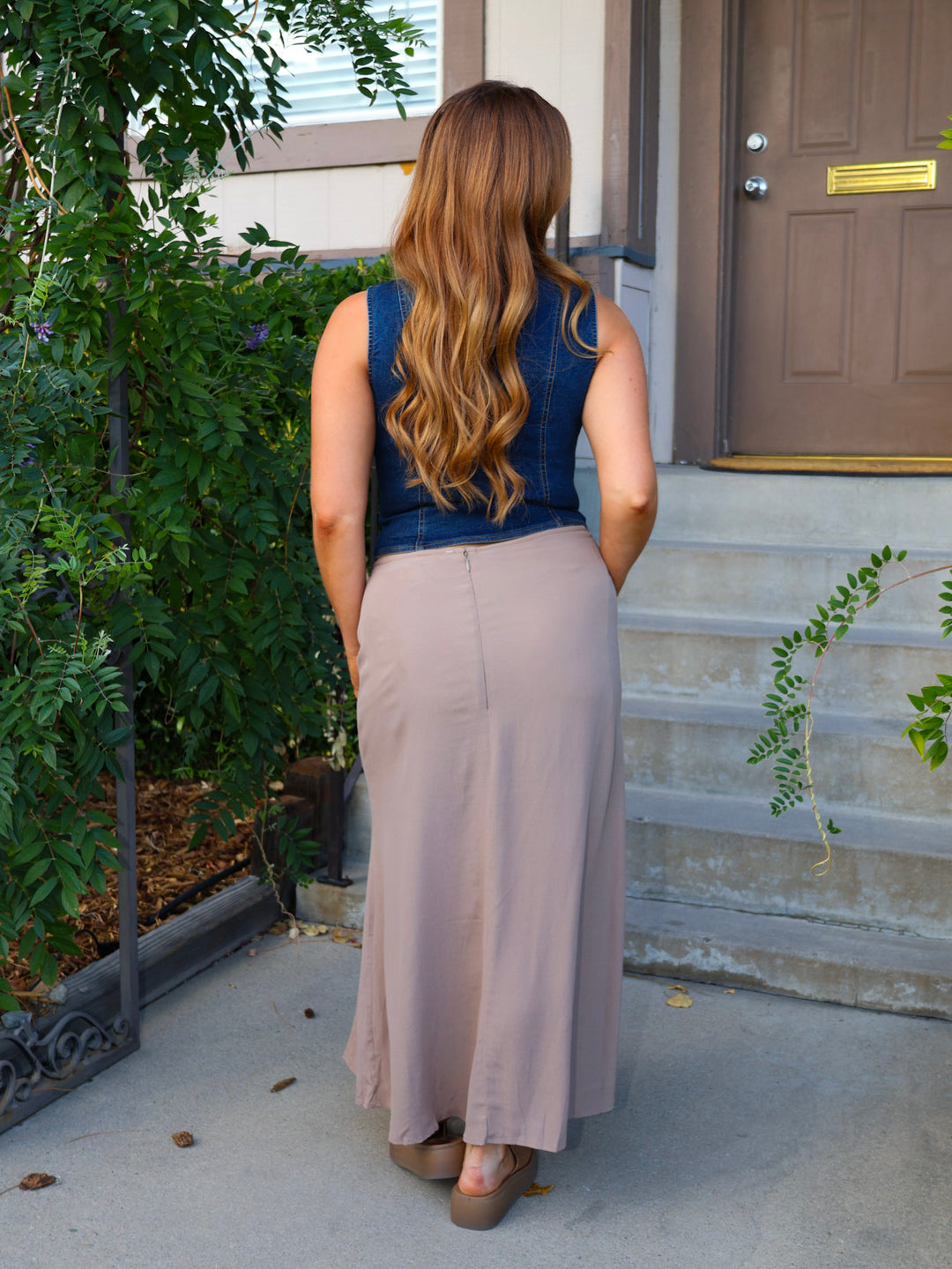 Woman standing on a porch wearing a denim top and beige skirt.