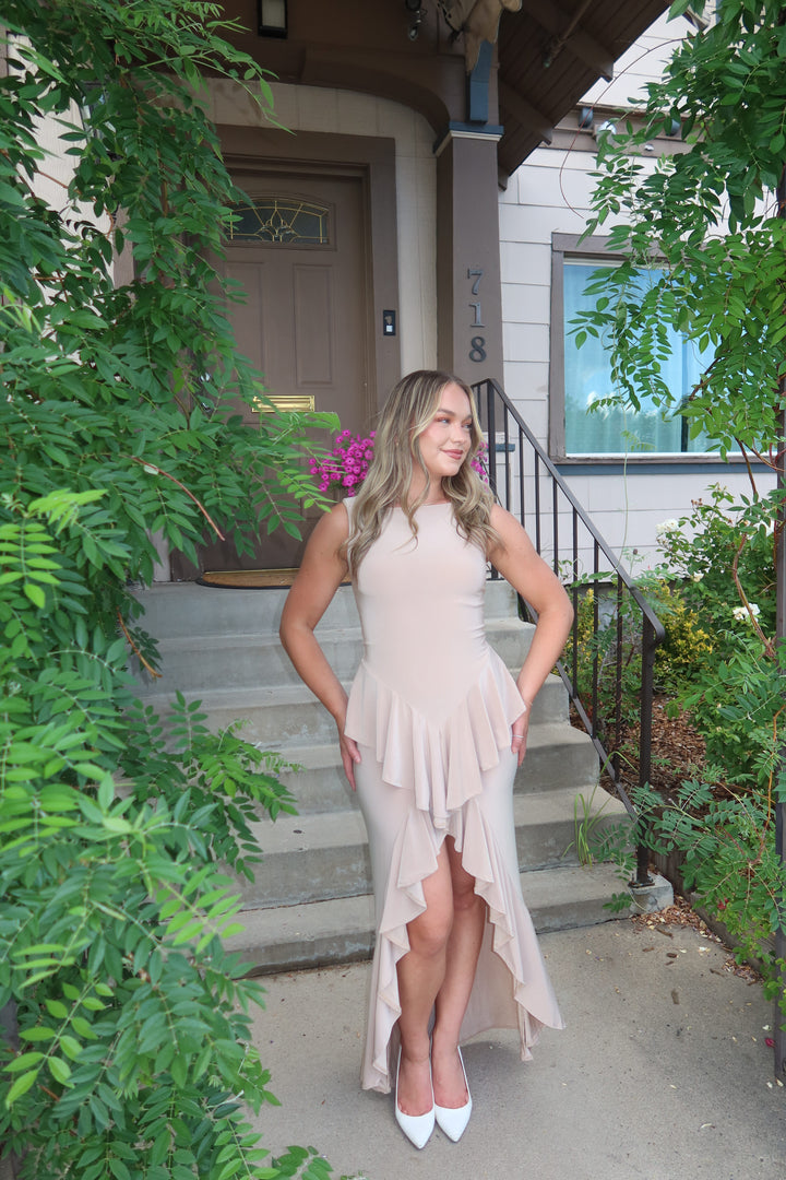 A woman wearing a sleeveless blush pink high-low ruffle dress and white pointed-toe heels stands on the steps of a house surrounded by green foliage.