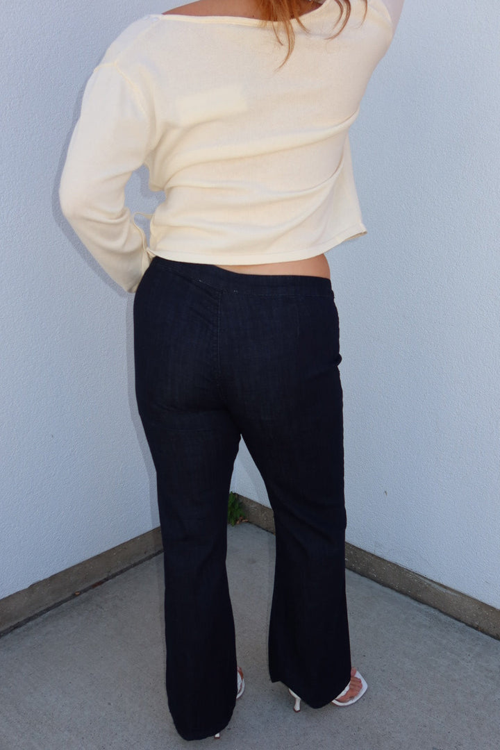 A person shown from the back wearing a cream long-sleeve cropped top and dark navy wide-leg jeans, standing in a corner against a light blue wall.