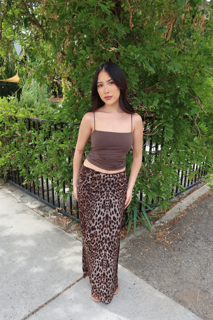 A woman stands outdoors on a sidewalk in front of green bushes and a black metal fence, wearing a brown ribbed camisole top and a brown leopard-print maxi skirt.