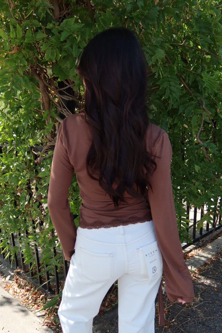 A woman standing outdoors on a sidewalk in front of green bushes, shown from behind. She is wearing a brown long-sleeve top and white wide-leg jeans.