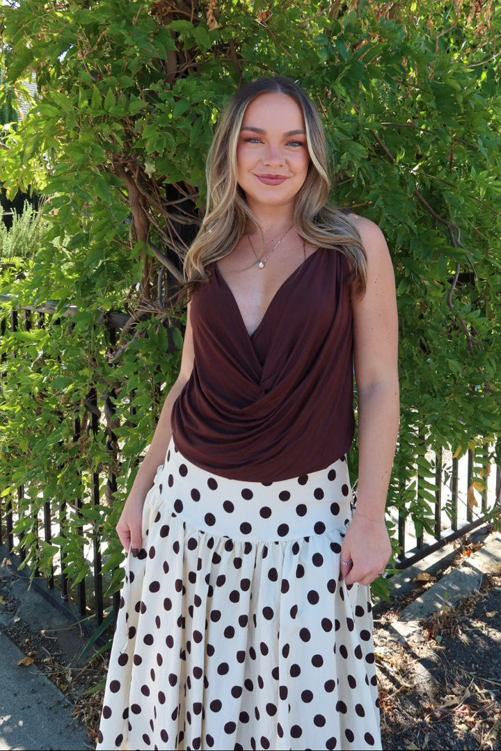 A woman standing outdoors in front of a green hedge and black metal fence, wearing a dark brown draped sleeveless top and a white skirt with large brown polka dots.