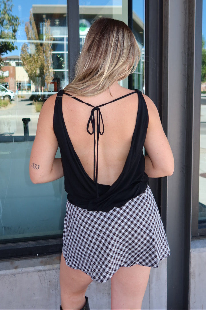 A woman viewed from behind wearing a black backless tie-front top and a black-and-white gingham mini skirt, standing in front of a storefront window.