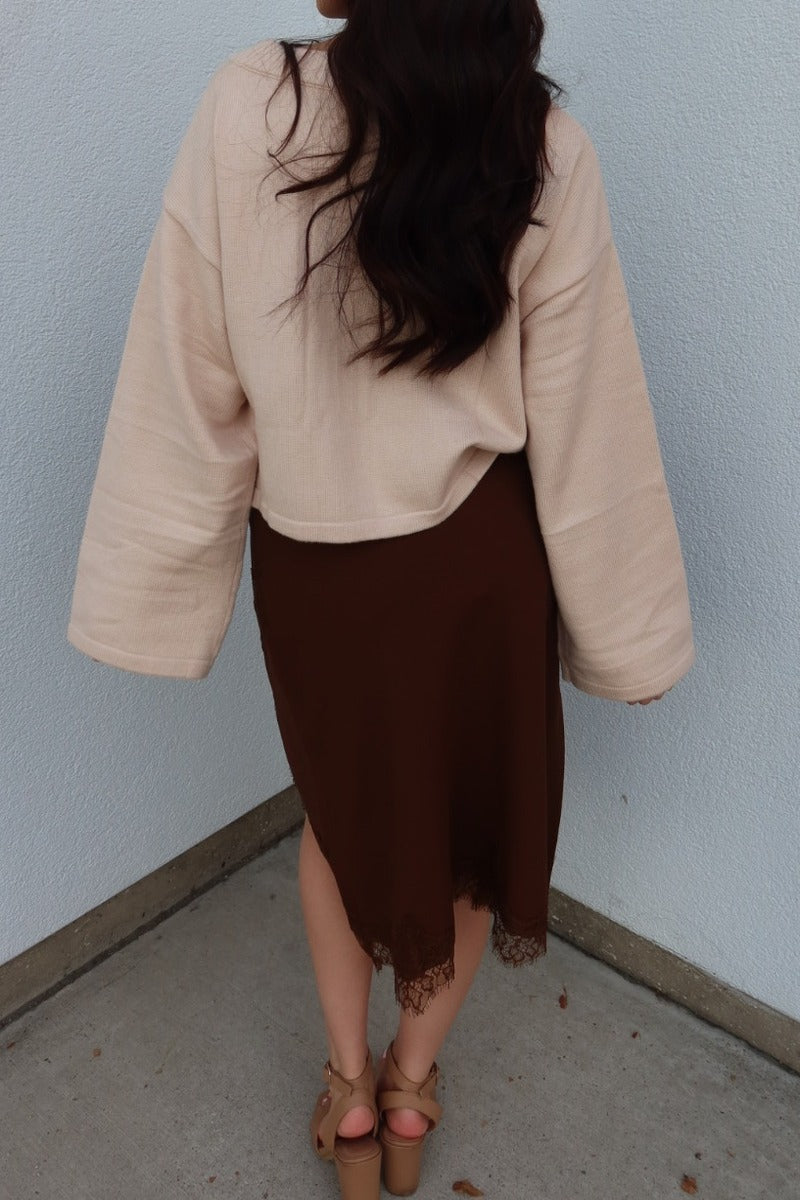 Back view of the brown Lucy asymmetric lace trim satin midi skirt worn with an oversized cream cardigan