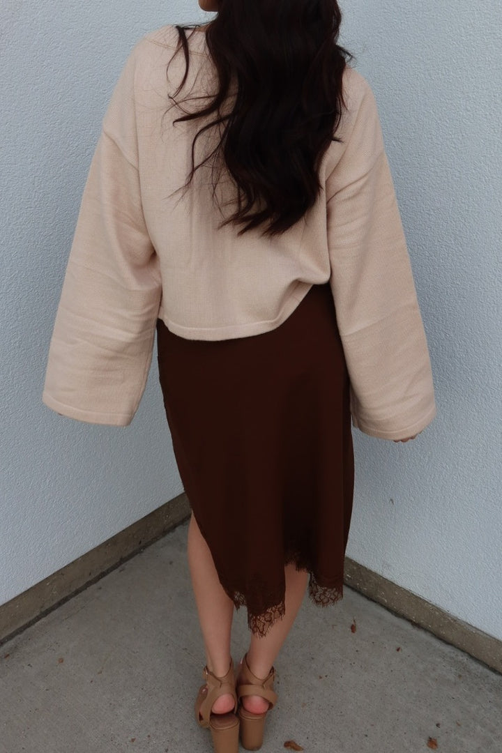 Back view of the brown Lucy asymmetric lace trim satin midi skirt worn with an oversized cream cardigan