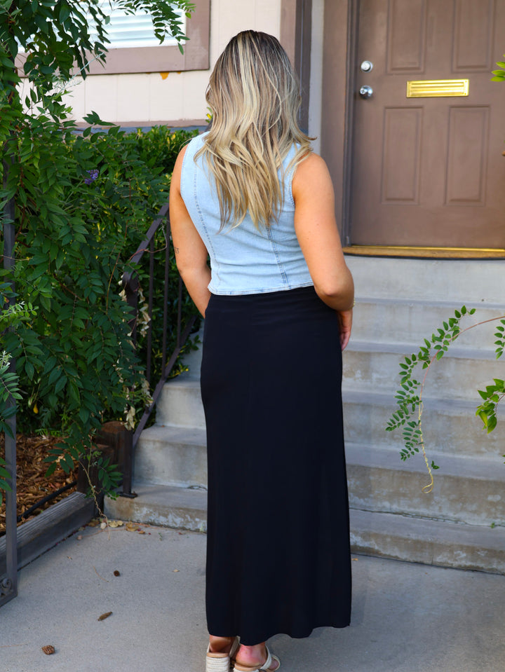 Woman standing outside a house wearing a light blue sleeveless top and black skirt.