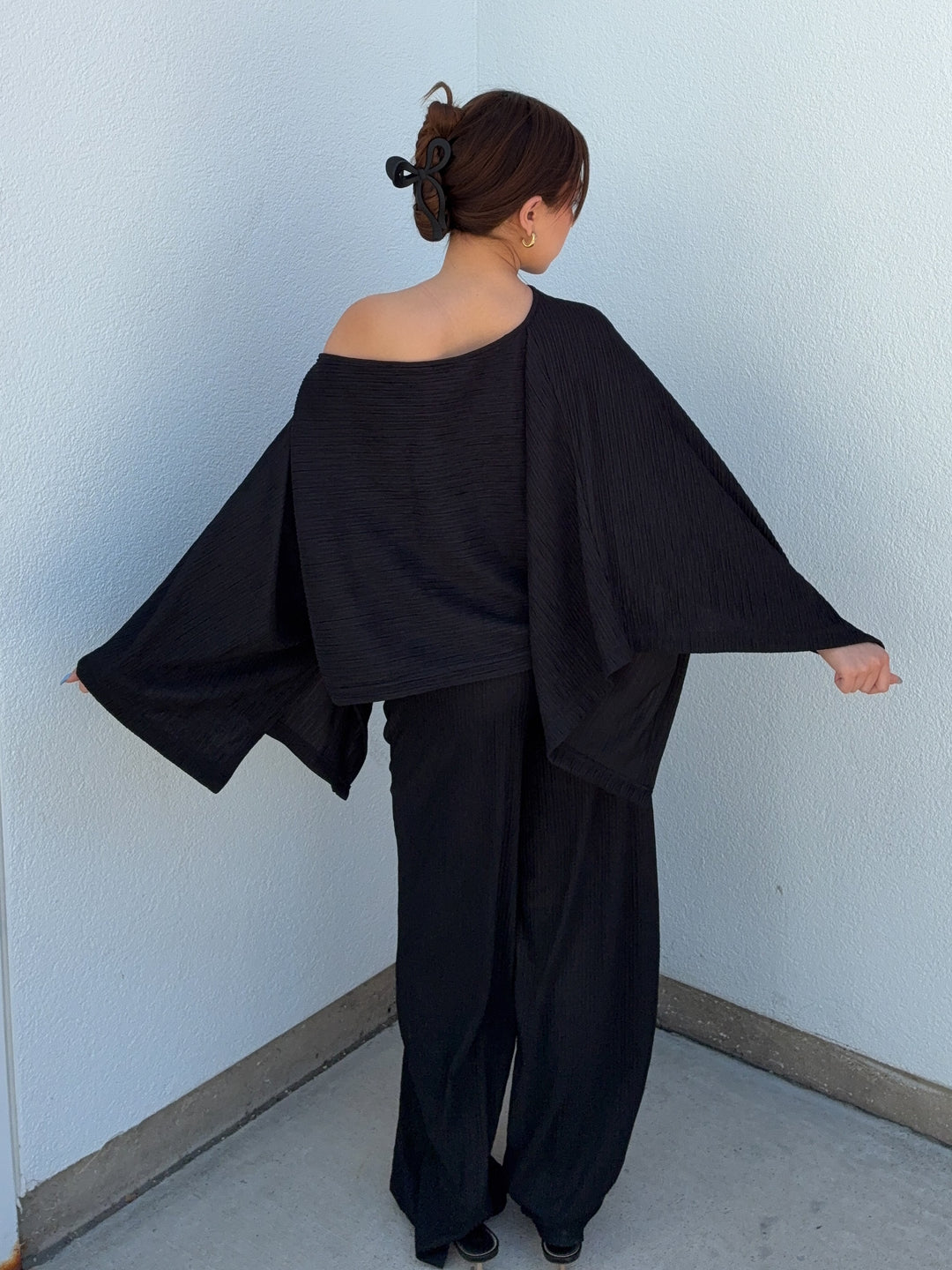 Person wearing a black outfit with wide sleeves against a light blue wall.