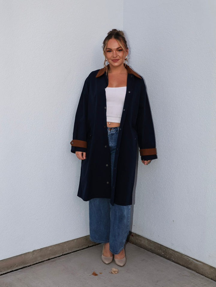 Woman wearing a long dark coat with brown accents against a white wall.