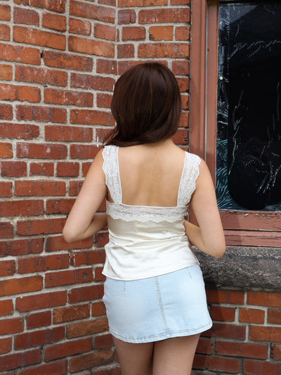 Woman standing against a brick wall wearing a white lace top and light blue denim skirt.