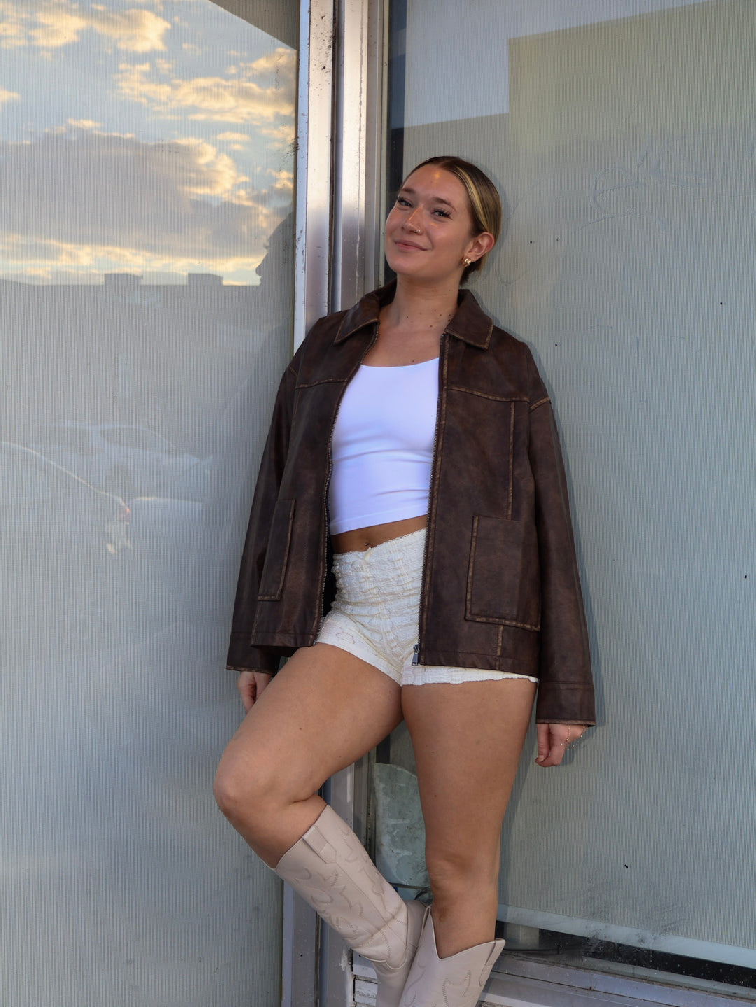 Woman wearing a brown jacket, white top, and shorts standing against a wall.