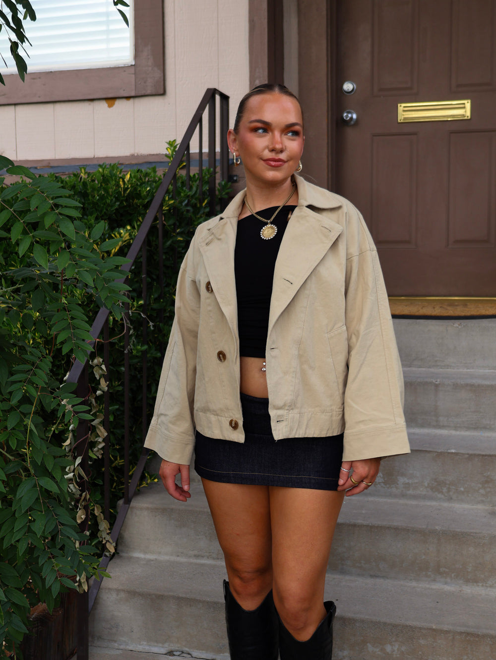 Woman wearing a beige jacket, black top, and skirt standing on steps outside a house.
