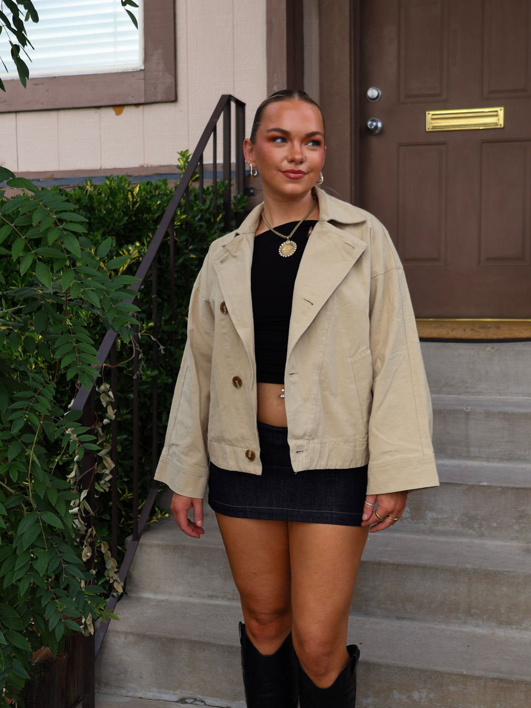 Woman wearing a beige jacket, black top, and skirt standing on steps outside a house.